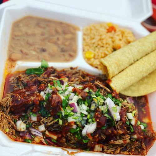 A takeout container with shredded beef in sauce, rice, beans, and three corn tortillas, garnished with onions and cilantro.