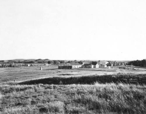 A black and white landscape showing a small settlement surrounded by grasslands and rolling hills in the background.