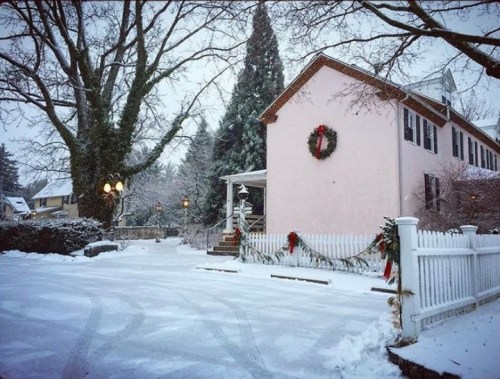 A snowy scene featuring a pink house adorned with a wreath, surrounded by trees and a white picket fence.