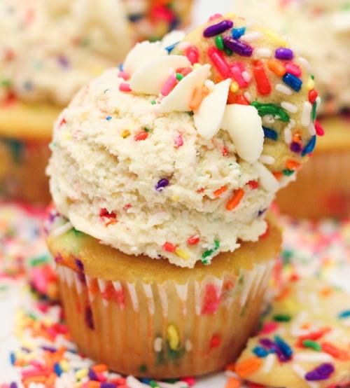 A colorful cupcake topped with frosting and sprinkles, surrounded by scattered sprinkles on a white surface.