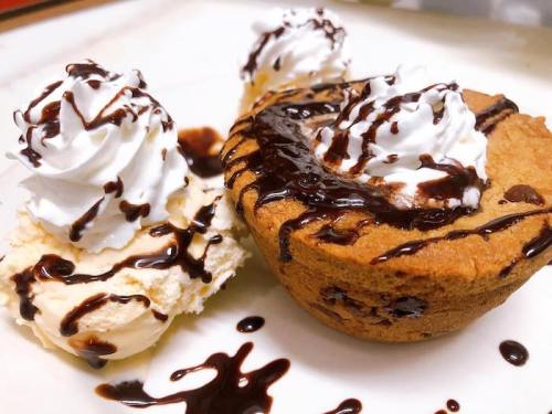 A warm chocolate chip cookie bowl topped with whipped cream and chocolate sauce, served with a scoop of vanilla ice cream.