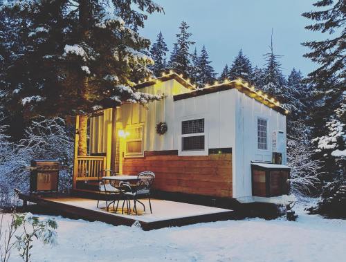 A cozy cabin in a snowy landscape, adorned with string lights, surrounded by trees. A small table and chairs are outside.