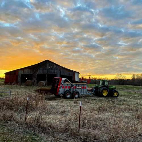 A tractor and trailer parked near a barn at sunset, with a colorful sky and grassy field in the foreground.