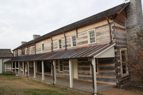 A historic log cabin with a metal roof, featuring multiple windows and a porch, set against a cloudy sky.