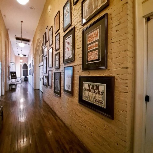 A hallway with wooden floors and brick walls, featuring framed posters and artwork on one side.
