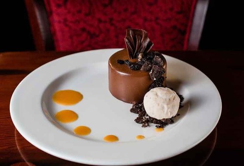 A chocolate dessert on a white plate, garnished with dark chocolate curls and a scoop of ice cream, with sauce drizzles.