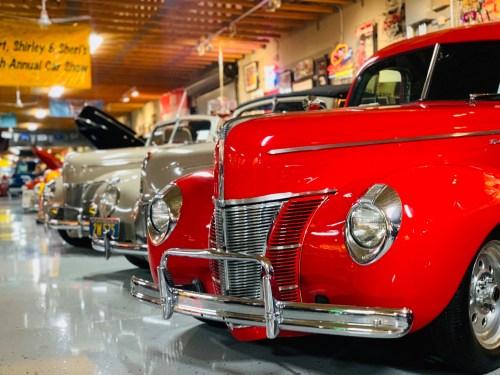 A row of classic cars, including a shiny red vintage model, displayed at a car show in a well-lit garage.