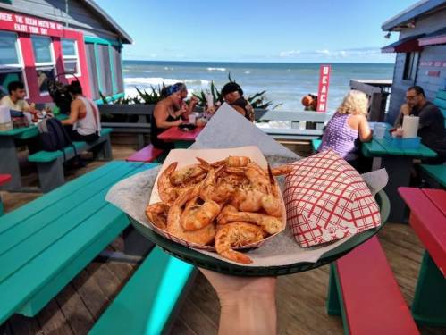 A hand holds a plate of shrimp in front of a beachside eatery with people dining outdoors.