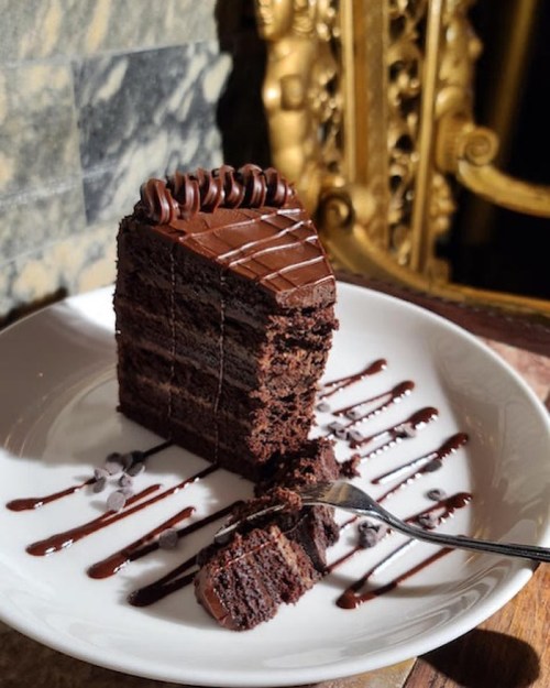 A slice of rich chocolate cake on a plate, drizzled with chocolate sauce and garnished with chocolate chips.