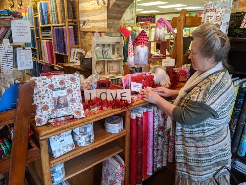 A woman arranges colorful fabric and craft supplies on a wooden display in a vibrant store filled with textiles.