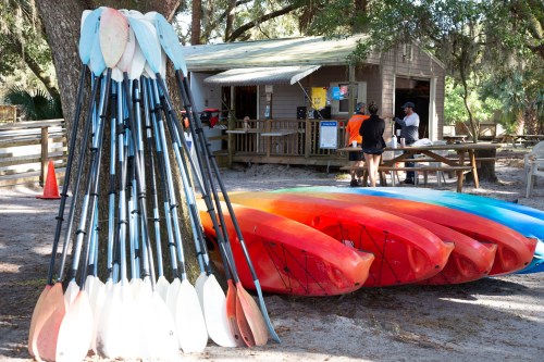 A stack of colorful kayaks and paddles near a rental hut in a shaded outdoor area.