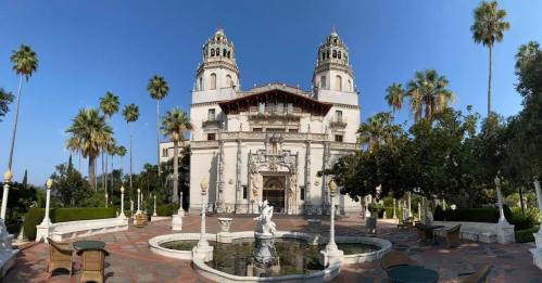 A grand mansion with two towers, surrounded by palm trees and a fountain in the foreground under a clear blue sky.