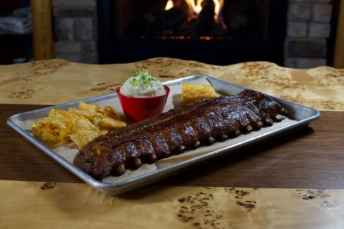 A platter with BBQ ribs, potato chips, and a small bowl of mashed potatoes, set against a cozy fireplace backdrop.