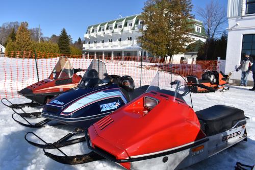 Three snowmobiles in bright colors parked on snow, with a hotel and trees in the background on a sunny day.