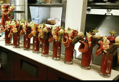 A row of tall glasses filled with a red drink, garnished with shrimp, vegetables, and various toppings.