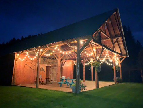 A rustic pavilion at night, illuminated by string lights, with a grassy area and picnic tables underneath.