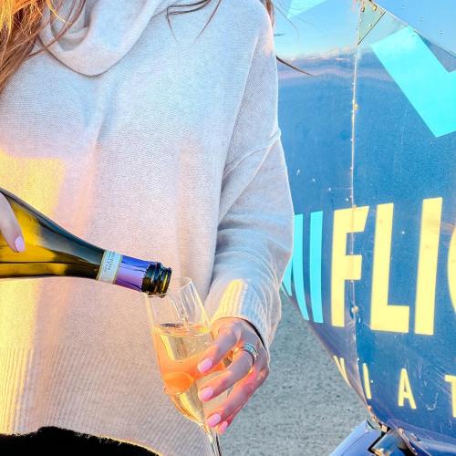 A person pours champagne into a glass beside a blue aircraft, with sunlight reflecting off the scene.