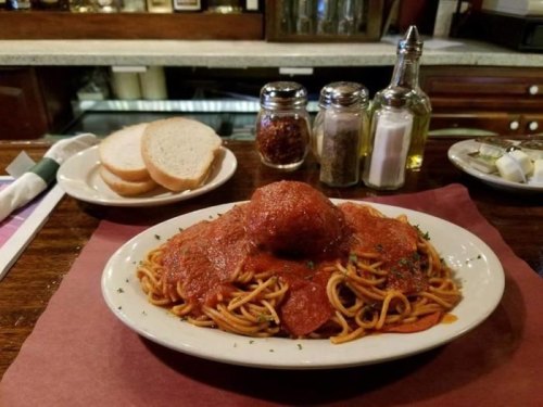 A plate of spaghetti topped with marinara sauce and a meatball, served with slices of bread and condiments in the background.