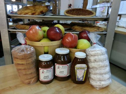 A display of baked goods, including pies, cookies, jars of apple butter, and fresh apples on a wooden table.
