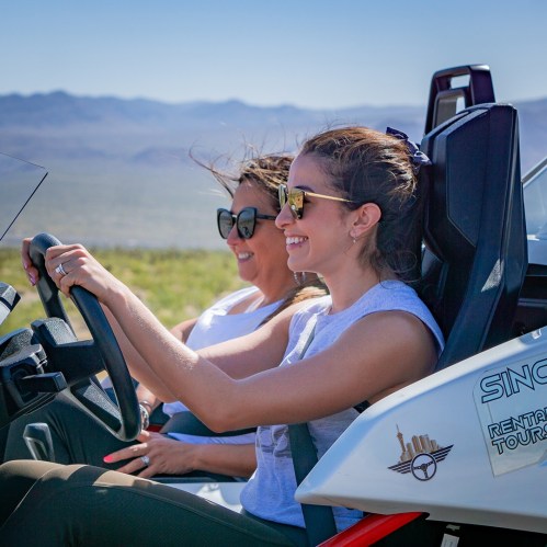 Two women smiling while driving a dune buggy in a scenic outdoor landscape.