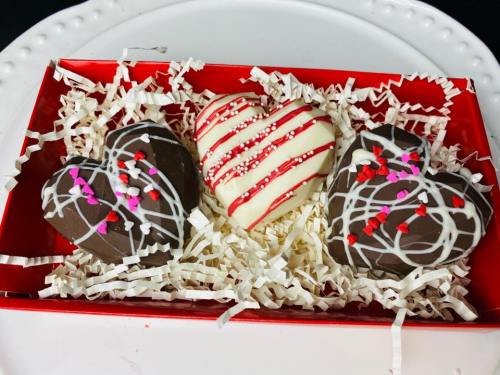 A red box filled with three heart-shaped chocolates decorated with white and pink icing and sprinkles on shredded paper.