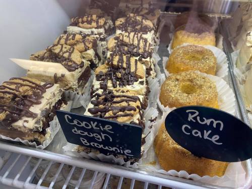 A display case with cookie dough brownies drizzled in chocolate and rum cakes, labeled on small signs.