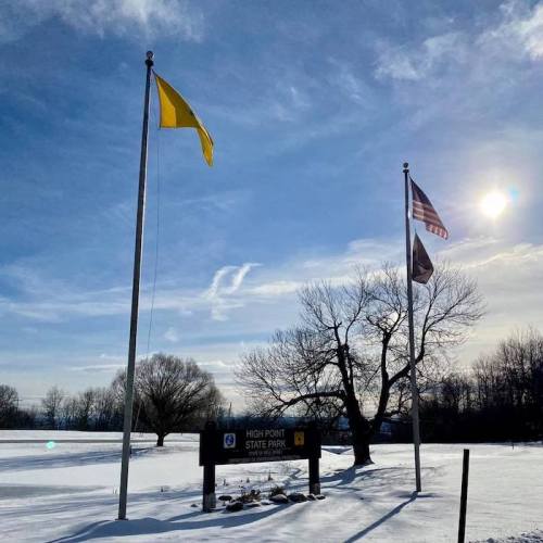 A snowy landscape at High Point State Park, featuring flags and a sign under a bright sky with scattered clouds.