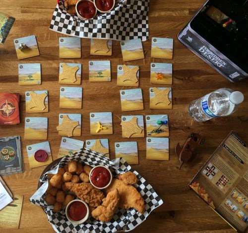 A tabletop game setup with food, cards, and a water bottle on a wooden surface.