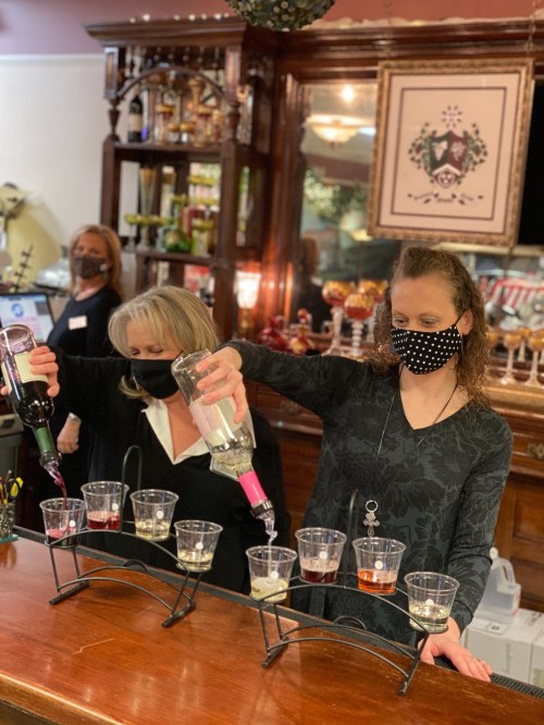 Two women in masks pour drinks into cups at a bar, with a decorative background and another person in the distance.
