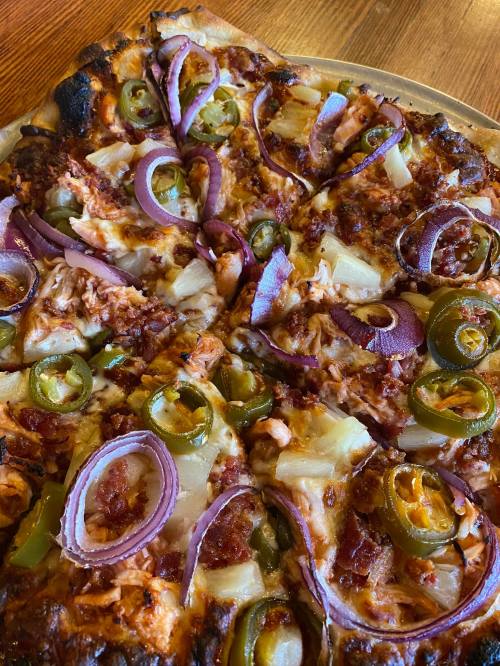 A close-up of a pizza topped with red onions, jalapeños, pineapple, and a mix of meats on a golden crust.