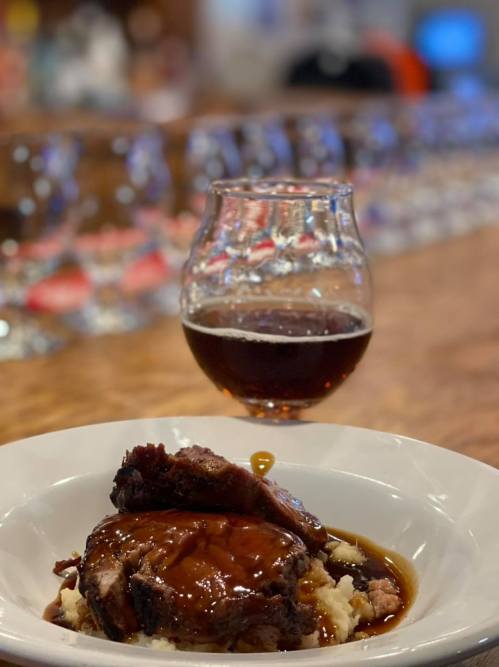 A plate of tender beef with gravy on mashed potatoes, accompanied by a glass of dark beer in a cozy setting.