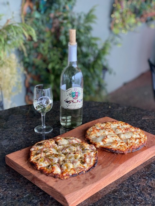 Two pizzas on a wooden board, one topped with vegetables, alongside a glass and bottle of white wine.