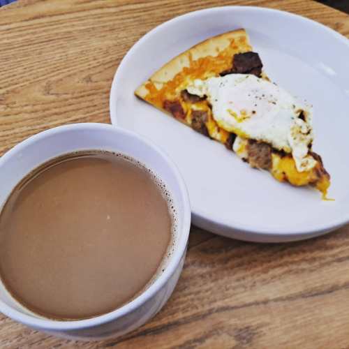 A slice of pizza topped with sausage and an egg, next to a cup of coffee on a wooden table.