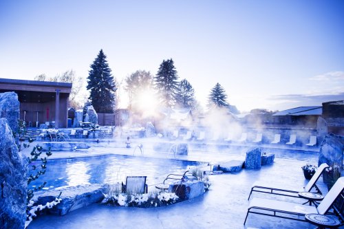 A serene outdoor hot spring pool surrounded by snow, with mist rising and sun shining through trees.