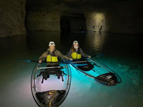 Two people in kayaks with headlamps, paddling in a dark cave with glowing water and distant figures in the background.