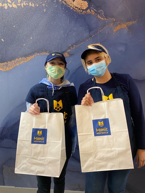 Two individuals wearing masks and hats hold white bags with a logo, standing in front of a blue and gold background.