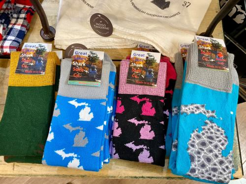 Colorful socks featuring Michigan shapes and shark designs, displayed on a wooden table with tags.