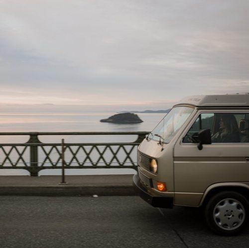 A vintage van drives along a bridge with a calm body of water and an island in the background under a cloudy sky.