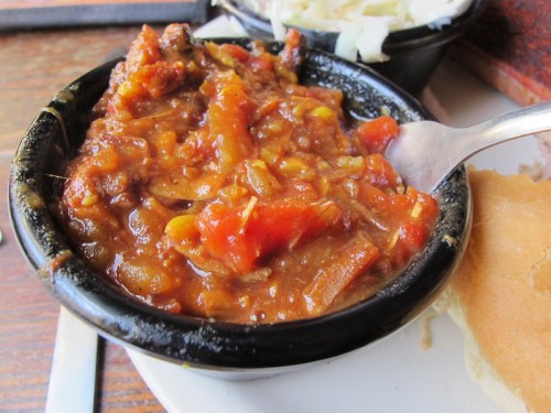 A bowl of chili with beans and tomatoes, served with a side of coleslaw and a bun on a plate.
