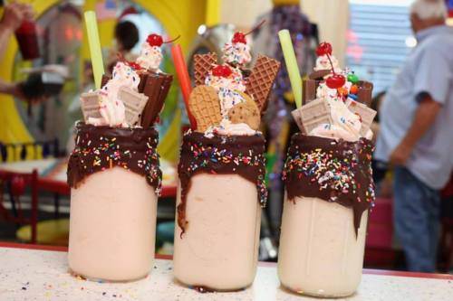 Three decadent milkshakes topped with whipped cream, chocolate, sprinkles, and various candies in colorful jars.