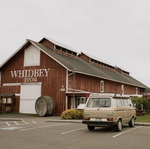 A vintage van parked in front of a large red barn with "WHIDBEY 1904" painted on it, surrounded by a cloudy sky.