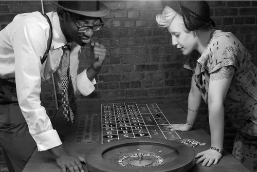 A man and woman lean over a roulette table, engaged in a game, with a vintage atmosphere and brick wall backdrop.