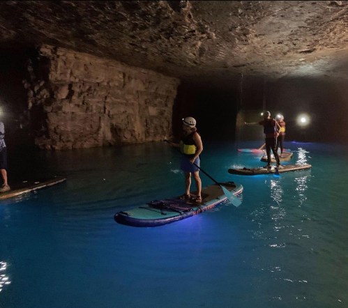People paddleboarding in a cave with glowing blue water, illuminated by headlamps in a dark, rocky environment.