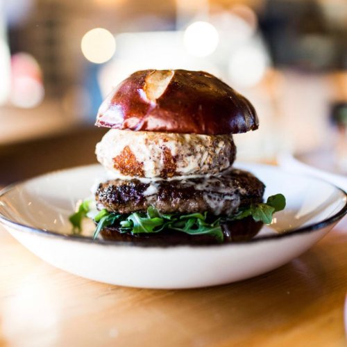 A gourmet burger with a pretzel bun, beef patty, crispy onion ring, and greens on a white plate.