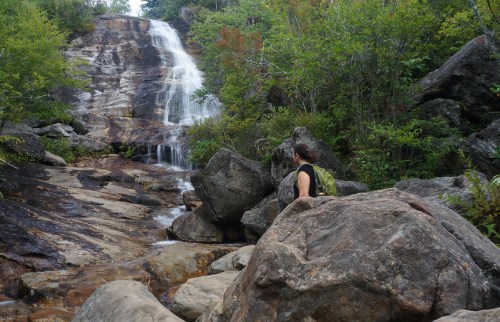 A person with a backpack gazes at a waterfall surrounded by lush greenery and rocky terrain.