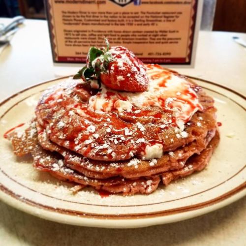 A stack of fluffy pancakes topped with whipped cream, strawberry, and syrup, served on a plate.