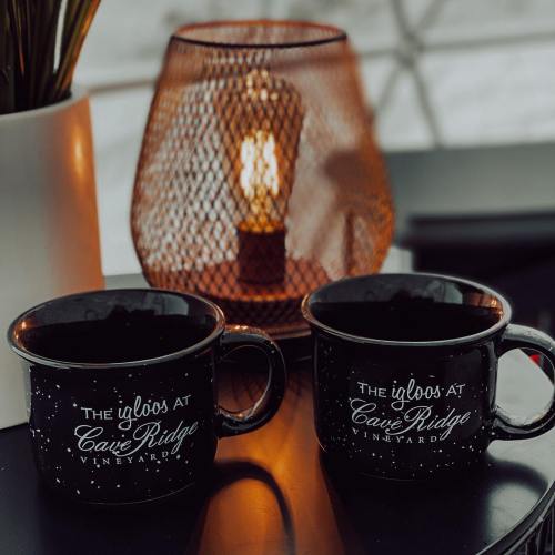 Two black mugs with "The Igloos at Cave Ridge Vineyard" text, placed on a table next to a warm candle in a mesh holder.