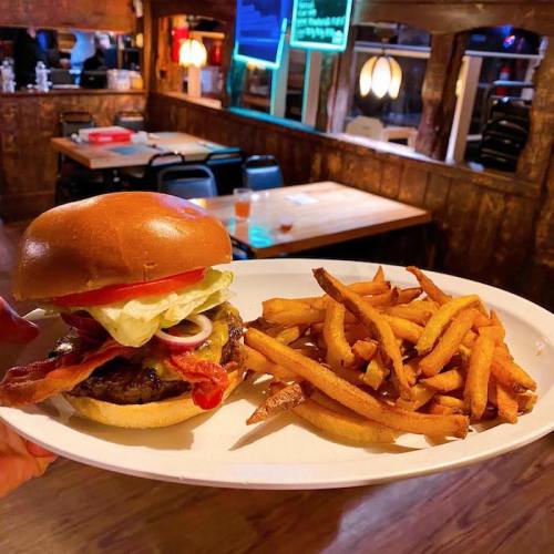 A hand holds a plate with a burger topped with lettuce, tomato, and bacon, alongside a serving of sweet potato fries.