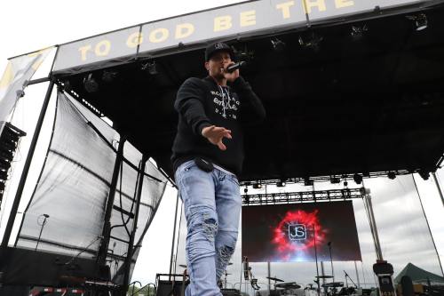 A performer raps on stage, wearing a black hoodie and distressed jeans, with a vibrant backdrop and sound equipment visible.