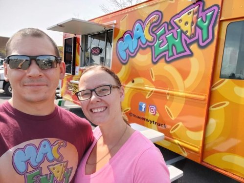 A couple poses in front of a colorful food truck with a cheese-themed design, promoting "Mac Envy."
